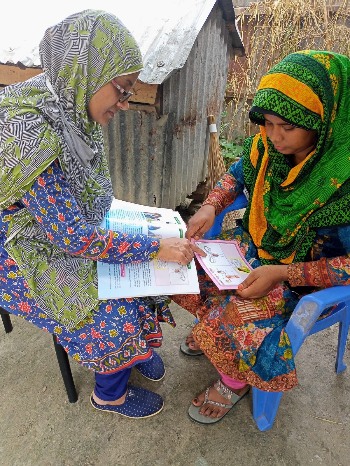 A Health Leader sits with a woman outside her home, working through a printed health education booklet together.