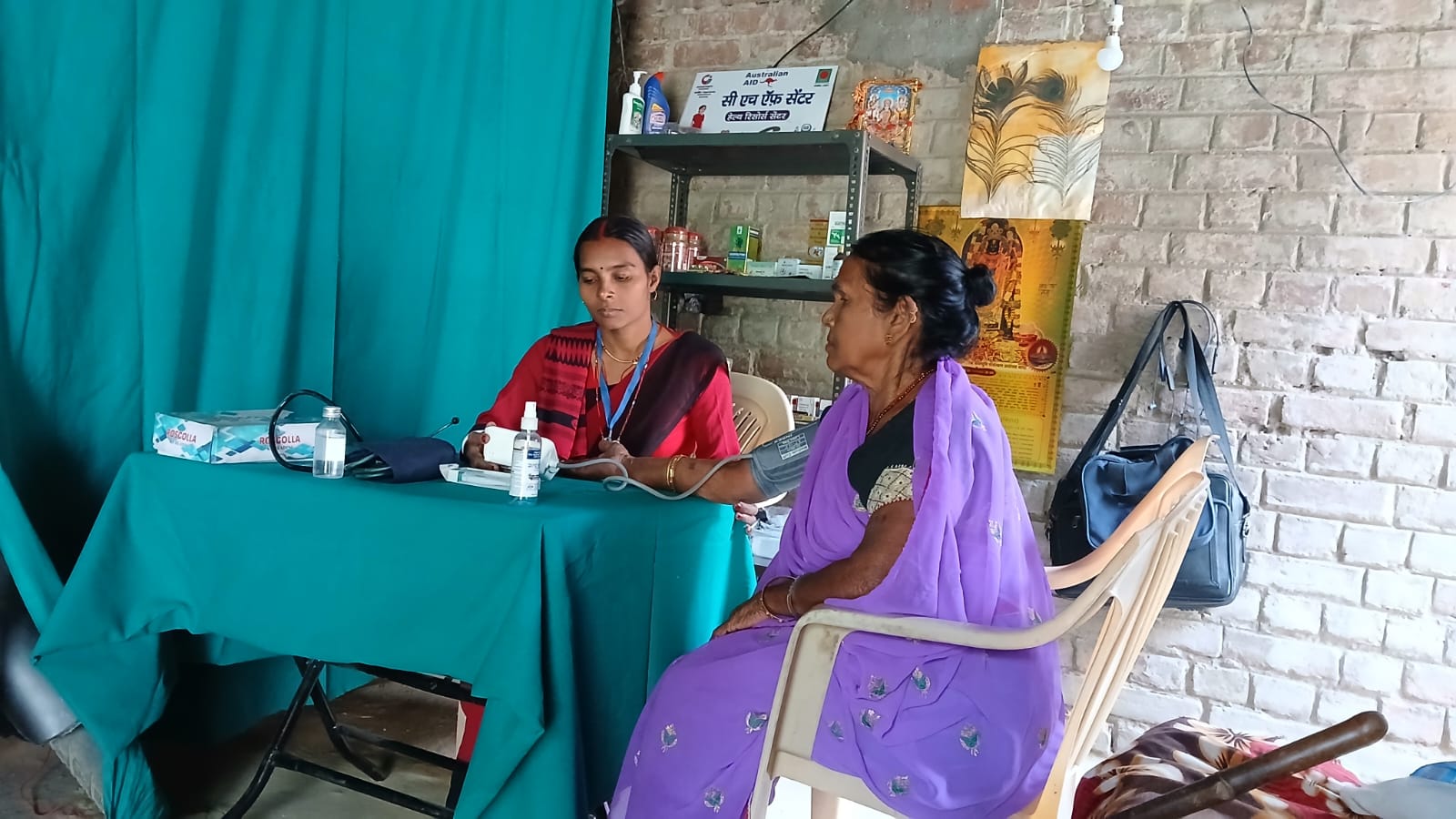A health worker takes a woman's blood pressure at a small community health centre, seated across a table from her.