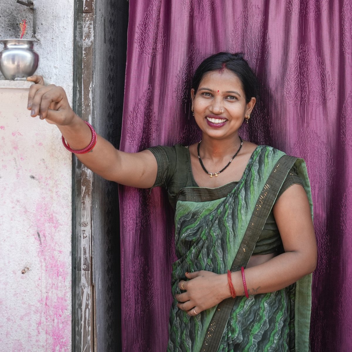 A woman in a green sari stands smiling in the doorway of her home, one hand resting on a shelf beside her.