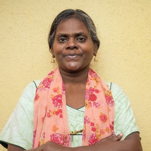 A woman stands against a plain wall, looking directly at the camera.