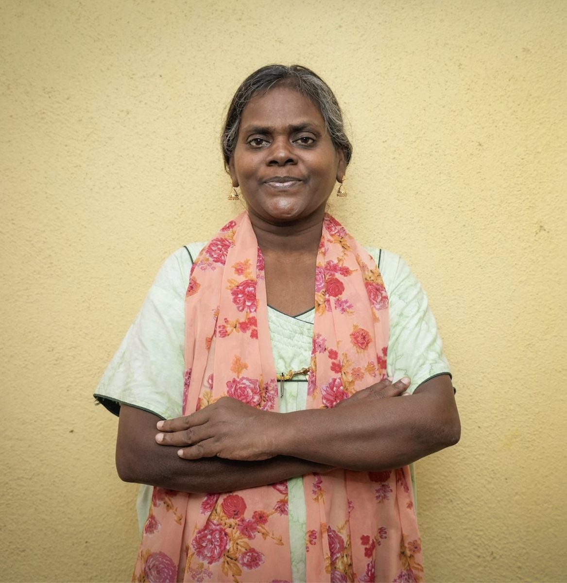 A woman stands against a plain wall with her arms folded, looking directly at the camera.