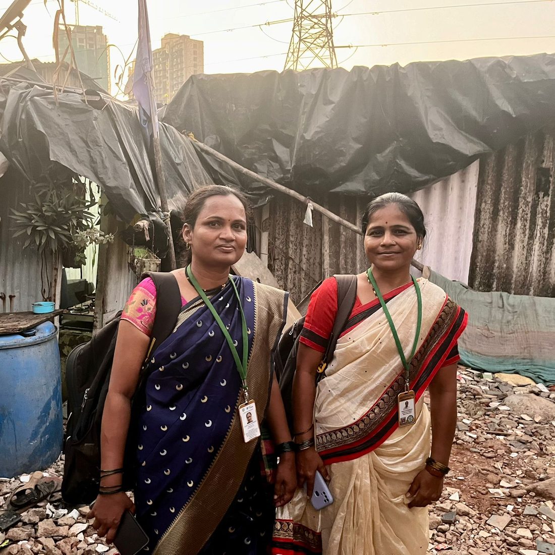 Two women wearing identification lanyards stand together in their community, with high-rise buildings in the distance.