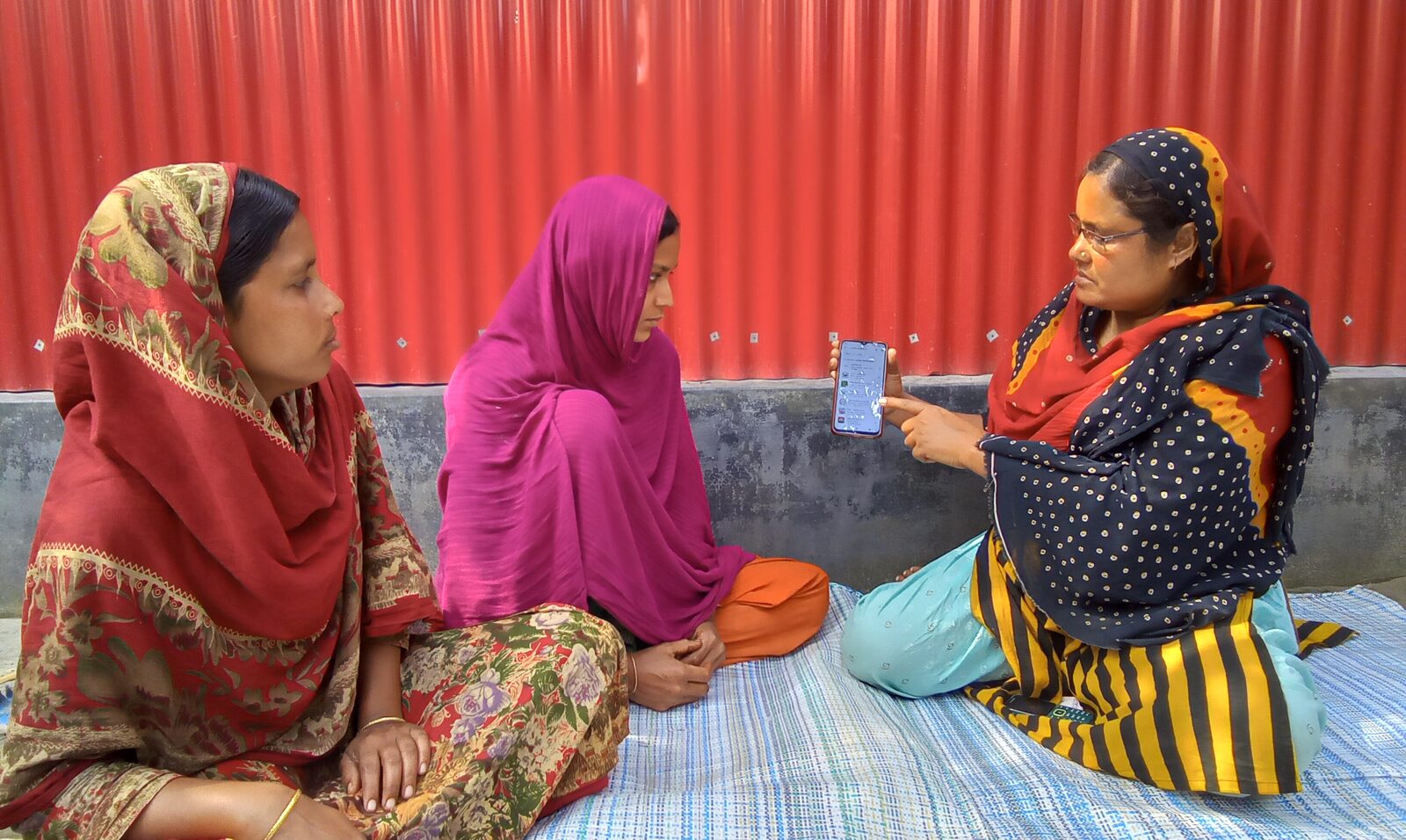 A Health Leader sits with two women on a mat, showing them a health consultation on her phone.