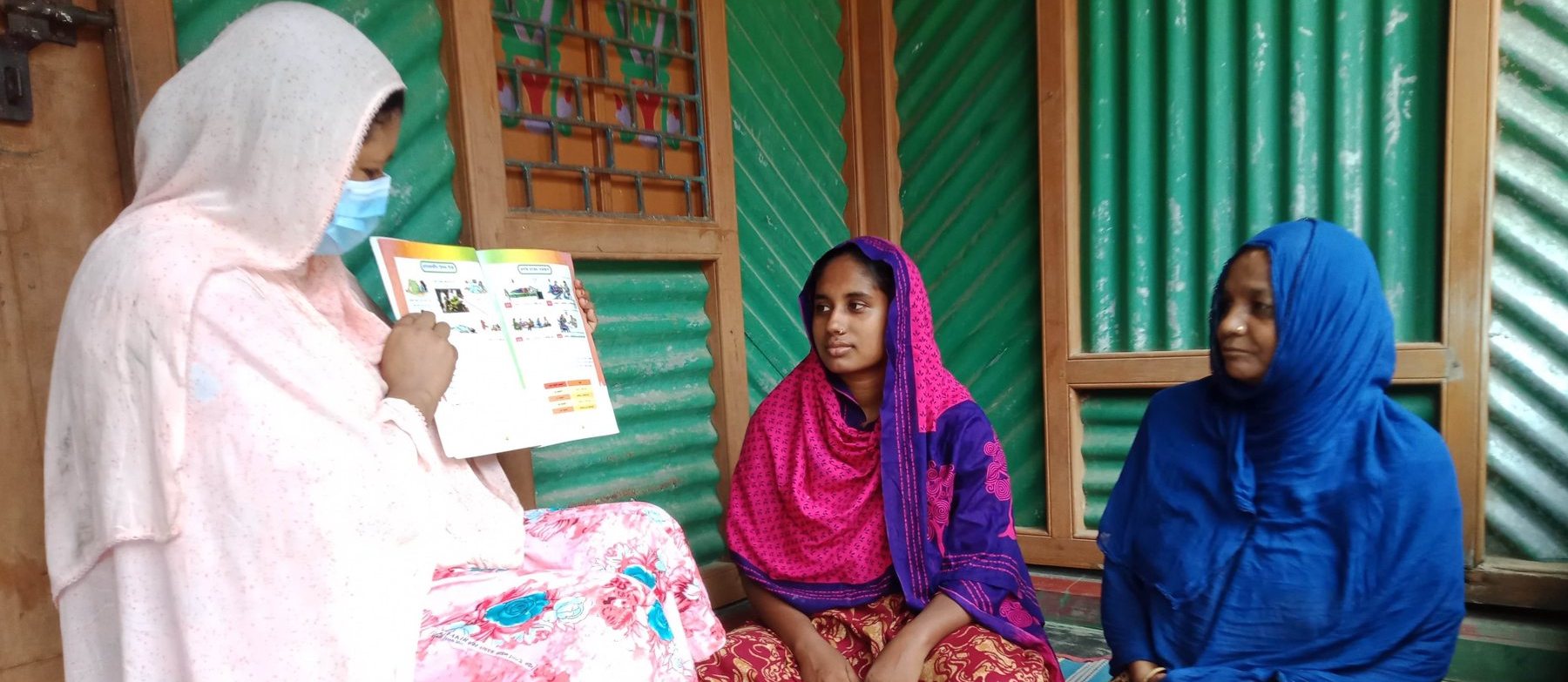 A Health Leader holds up an illustrated health education booklet for two women seated in front of her.
