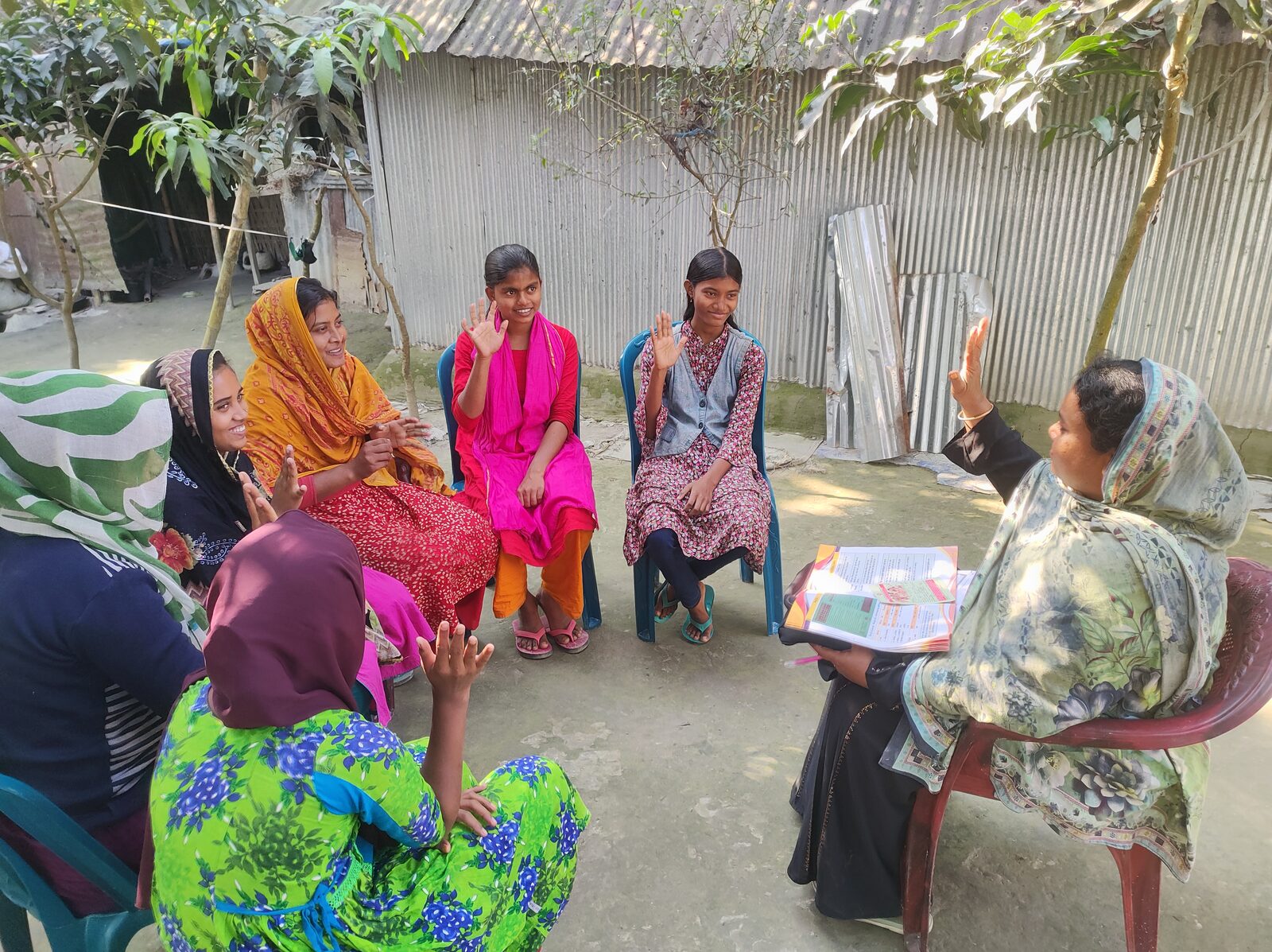 A Health Leader leads an outdoor session with a group of young women and adolescent girls seated in a circle, some with hands raised.