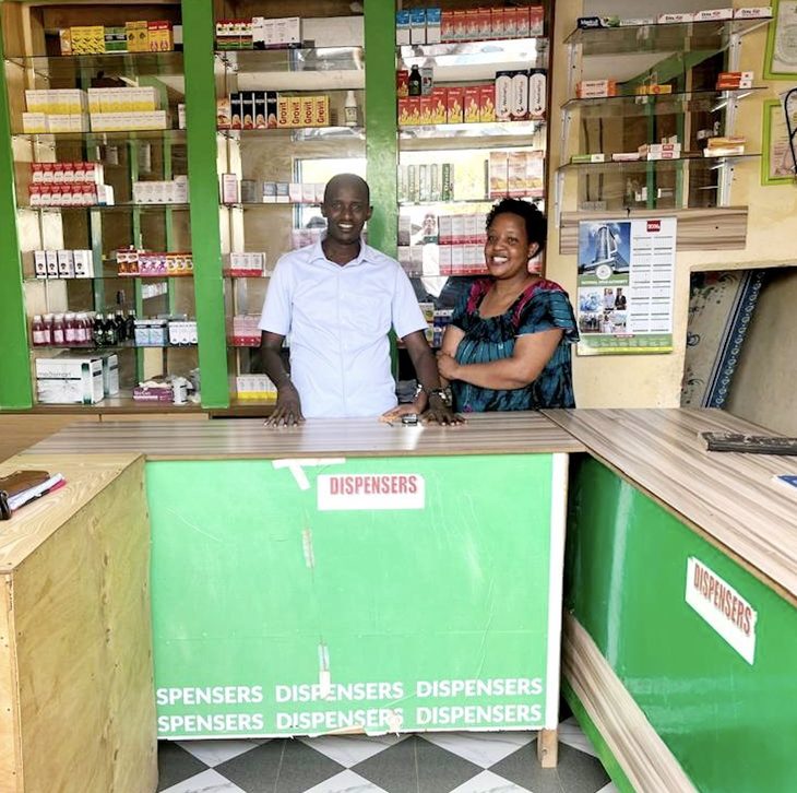 Two pharmacy owners stand behind the counter of their community pharmacy, shelves of medicines stocked behind them.