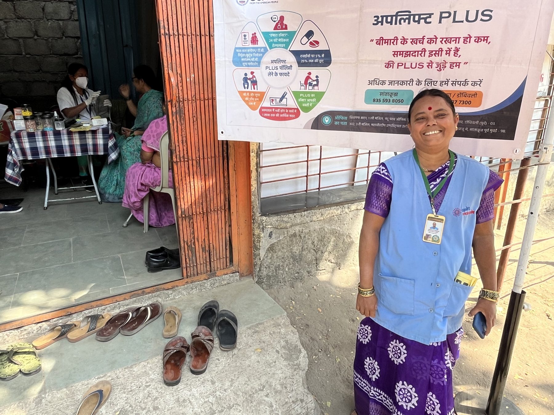 A health worker in program uniform stands beside a banner advertising a community health insurance policy, with a health camp running behind her.