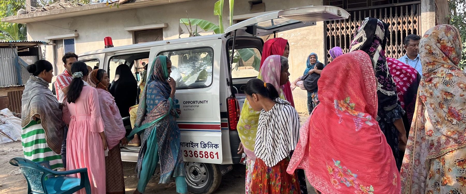 Women wait beside a mobile health clinic vehicle with its rear door open, outside houses in a rural community.