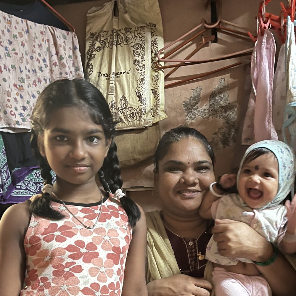 A woman sits holding her baby, with her older daughter standing beside her, inside their home.
